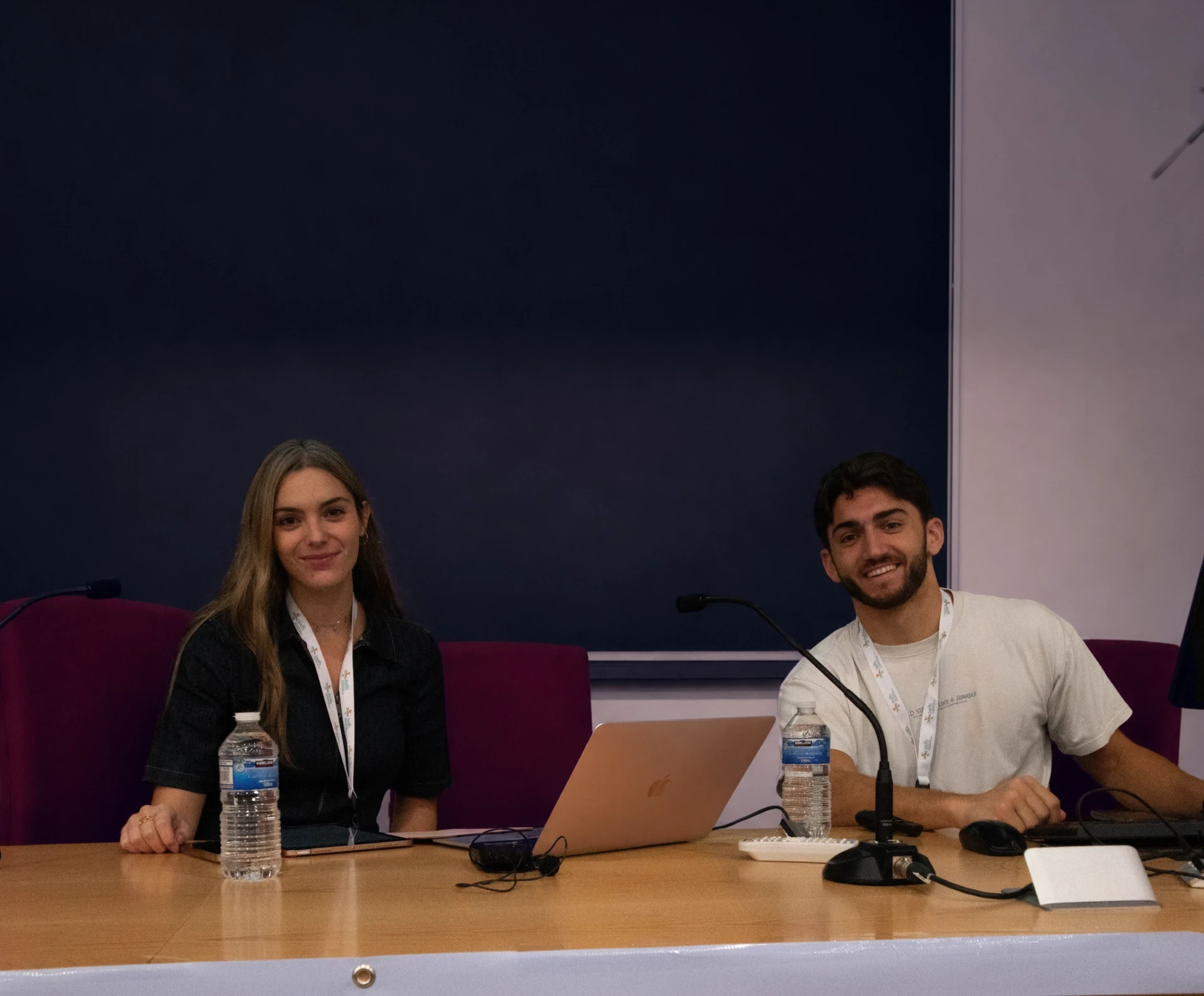 Dos personas sentadas a una mesa de ponentes con micrófonos y un portátil abierto, sonriendo a cámara durante una jornada de formación