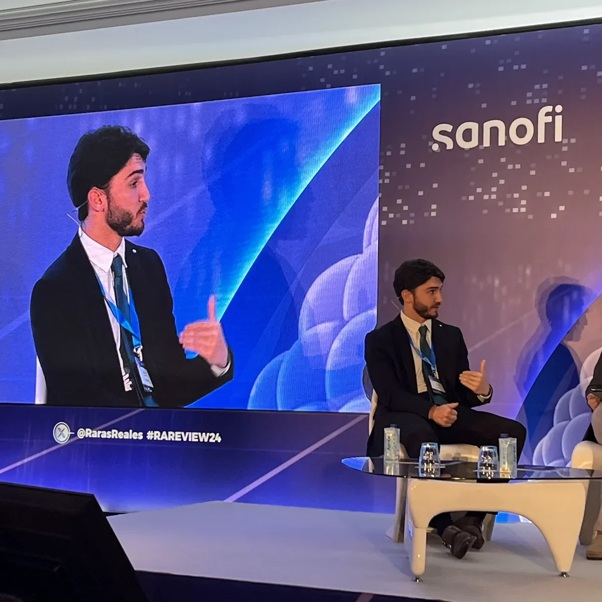 Un ponente sentado en el escenario de un panel de Sanofi, con su plano corto proyectado a tamaño gigante detrás